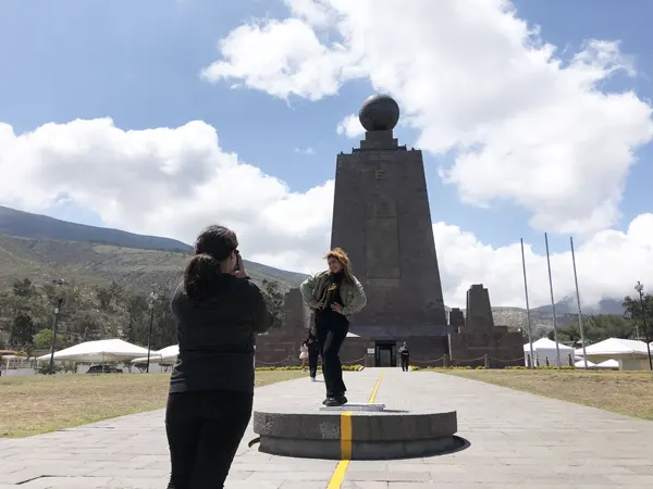 One girl taking a picture of another on the Equator line with the monument in the background